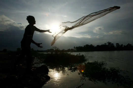 A man tosses a fishing net into a body of water with the sun rising in the background.
