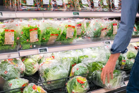 A person's arm reaches for a package of greens from a selction of greens and lettuces in a grocery story.